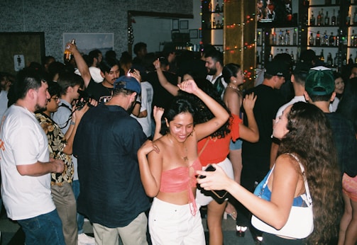 A lively scene at a crowded party with people dancing and socializing. The dimly lit room is adorned with colorful lights, and the background features shelves filled with various bottles. The attendees are dressed casually, enjoying the energetic atmosphere.