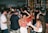 A lively scene at a crowded party with people dancing and socializing. The dimly lit room is adorned with colorful lights, and the background features shelves filled with various bottles. The attendees are dressed casually, enjoying the energetic atmosphere.