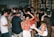 A lively scene at a crowded party with people dancing and socializing. The dimly lit room is adorned with colorful lights, and the background features shelves filled with various bottles. The attendees are dressed casually, enjoying the energetic atmosphere.