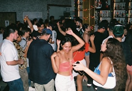 A lively scene at a crowded party with people dancing and socializing. The dimly lit room is adorned with colorful lights, and the background features shelves filled with various bottles. The attendees are dressed casually, enjoying the energetic atmosphere.