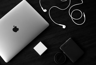 A silver-colored laptop with an Apple logo sits on a dark surface next to a pair of white earphones. Nearby, there is a small white rectangular object and a black external hard drive with a camera lens cap. The composition is minimalistic, with a monochrome aesthetic emphasizing the contrast between the objects and the background.