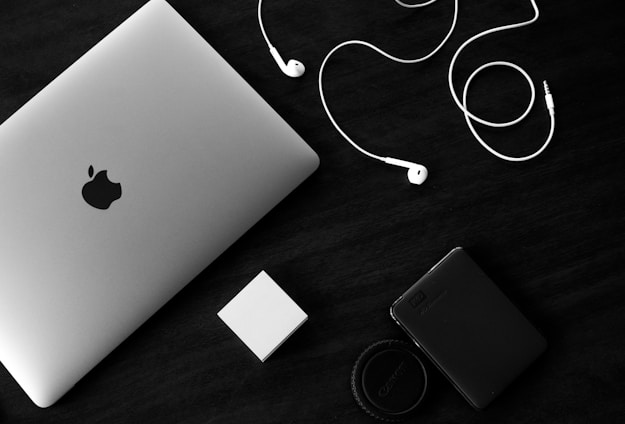 A silver-colored laptop with an Apple logo sits on a dark surface next to a pair of white earphones. Nearby, there is a small white rectangular object and a black external hard drive with a camera lens cap. The composition is minimalistic, with a monochrome aesthetic emphasizing the contrast between the objects and the background.