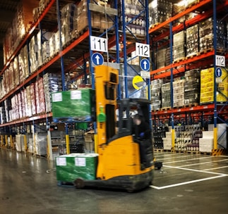 A bustling warehouse environment with stacked shelves filled with a variety of boxed goods. A compact forklift is actively moving, carrying wrapped pallets of green boxes. The shelves are organized with numbered signs, and the scene is well-lit, indicating a busy and efficient setting.