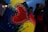 A heart-shaped design is featured prominently, with the words 'UKRAINE FOREVER' written inside. The heart is surrounded by a colorful artistic background with hues of blue, yellow, and red. The background includes sparkles and swirling brush strokes. In the dimly lit scene, there are blurred figures in the background, suggesting a night-time setting, possibly during a public gathering or protest.