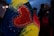 A heart-shaped design is featured prominently, with the words 'UKRAINE FOREVER' written inside. The heart is surrounded by a colorful artistic background with hues of blue, yellow, and red. The background includes sparkles and swirling brush strokes. In the dimly lit scene, there are blurred figures in the background, suggesting a night-time setting, possibly during a public gathering or protest.