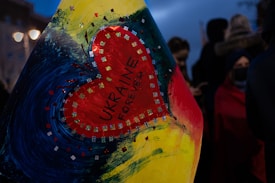A heart-shaped design is featured prominently, with the words 'UKRAINE FOREVER' written inside. The heart is surrounded by a colorful artistic background with hues of blue, yellow, and red. The background includes sparkles and swirling brush strokes. In the dimly lit scene, there are blurred figures in the background, suggesting a night-time setting, possibly during a public gathering or protest.