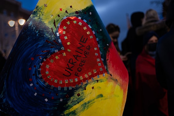 A heart-shaped design is featured prominently, with the words 'UKRAINE FOREVER' written inside. The heart is surrounded by a colorful artistic background with hues of blue, yellow, and red. The background includes sparkles and swirling brush strokes. In the dimly lit scene, there are blurred figures in the background, suggesting a night-time setting, possibly during a public gathering or protest.