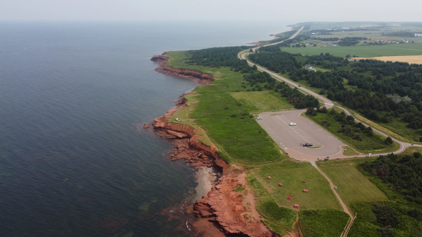 Aerial view of Prince Edward Island's red sandstone cliffs meeting the Gulf of St. Lawrence, green farmland and forest stretching inland from the eroded coastal edge