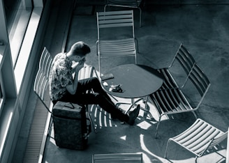 A person is sitting alone at a round table surrounded by empty chairs in what appears to be a public space. They have a suitcase next to them and are engaged with a smartphone placed on the table. Overhead lighting creates shadows on the floor, enhancing the solitude of the scene.