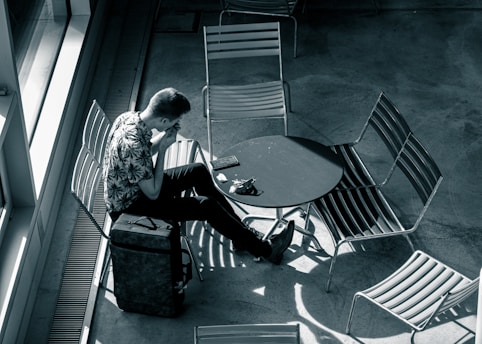 A person is sitting alone at a round table surrounded by empty chairs in what appears to be a public space. They have a suitcase next to them and are engaged with a smartphone placed on the table. Overhead lighting creates shadows on the floor, enhancing the solitude of the scene.
