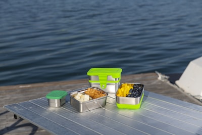 Several stainless steel food containers with green lids are placed on a metallic table, overlooking a body of water. The containers are filled with assorted fruits and snacks, including blueberries, mango slices, and pieces of bread or chips. The scene suggests a leisurely outdoor meal or picnic by the water.