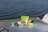 Several stainless steel food containers with green lids are placed on a metallic table, overlooking a body of water. The containers are filled with assorted fruits and snacks, including blueberries, mango slices, and pieces of bread or chips. The scene suggests a leisurely outdoor meal or picnic by the water.