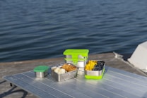 Several stainless steel food containers with green lids are placed on a metallic table, overlooking a body of water. The containers are filled with assorted fruits and snacks, including blueberries, mango slices, and pieces of bread or chips. The scene suggests a leisurely outdoor meal or picnic by the water.