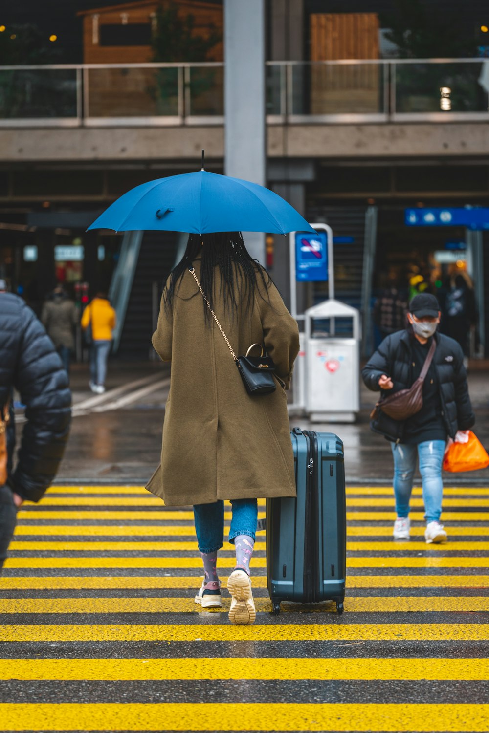 a woman walking across a cross walk holding a blue umbrella