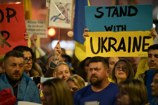A group of people holding signs and banners in support of Ukraine. The central sign has bold letters saying 'Stand with Ukraine.' There are various other signs and flags visible, with people of diverse backgrounds participating in the gathering.