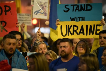 A group of people holding signs and banners in support of Ukraine. The central sign has bold letters saying 'Stand with Ukraine.' There are various other signs and flags visible, with people of diverse backgrounds participating in the gathering.