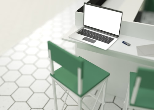 A laptop with a blank screen is placed on a counter with a USB flash drive and a closed laptop beside it. The setting includes two green barstools and a floor with hexagonal tiles in a white and light gray pattern.