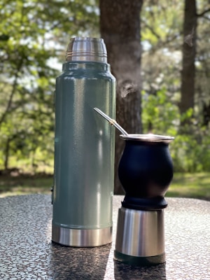 A tall thermos and a smaller cup with a metal straw are placed on a textured outdoor table. Steam is rising from the cup, suggesting a hot beverage. The background consists of blurred greenery and trees, indicating a setting in nature.