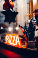 A dimly lit window display features a neon sign with the word 'PIZZA' glowing in bright orange. Next to the sign, a potted plant with long, pointed leaves adds a touch of greenery. Reflections on the glass and a hanging black and red star enhance the cozy and inviting ambiance.