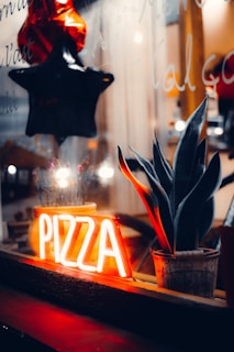 A dimly lit window display features a neon sign with the word 'PIZZA' glowing in bright orange. Next to the sign, a potted plant with long, pointed leaves adds a touch of greenery. Reflections on the glass and a hanging black and red star enhance the cozy and inviting ambiance.