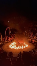 A dramatic scene featuring a circular arena with a central fire. Performers surround the flames, with an audience seated in an amphitheater fashion. The atmosphere is intense, highlighted by the darkness around and the bright orange of the flames.