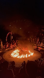 A dramatic scene featuring a circular arena with a central fire. Performers surround the flames, with an audience seated in an amphitheater fashion. The atmosphere is intense, highlighted by the darkness around and the bright orange of the flames.