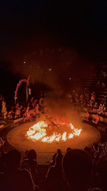 A dramatic scene featuring a circular arena with a central fire. Performers surround the flames, with an audience seated in an amphitheater fashion. The atmosphere is intense, highlighted by the darkness around and the bright orange of the flames.