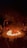 A dramatic scene featuring a circular arena with a central fire. Performers surround the flames, with an audience seated in an amphitheater fashion. The atmosphere is intense, highlighted by the darkness around and the bright orange of the flames.