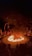 A dramatic scene featuring a circular arena with a central fire. Performers surround the flames, with an audience seated in an amphitheater fashion. The atmosphere is intense, highlighted by the darkness around and the bright orange of the flames.