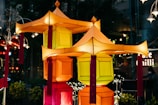Colorful lanterns with geometric shapes and tassels are prominently displayed against a dimly lit background. The lanterns exhibit vibrant hues of orange, yellow, and red, creating a festive and warm atmosphere. They are part of a decorative arrangement resembling traditional Asian designs.