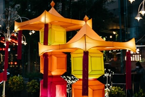 Colorful lanterns with geometric shapes and tassels are prominently displayed against a dimly lit background. The lanterns exhibit vibrant hues of orange, yellow, and red, creating a festive and warm atmosphere. They are part of a decorative arrangement resembling traditional Asian designs.