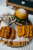 A round wooden tray holds a variety of foods including skewered meat or tofu, a burger with a powdered bun, and a glass dish filled with what appears to be a mushroom or popcorn-like snack. A blackboard with white letters is partially visible in the background.