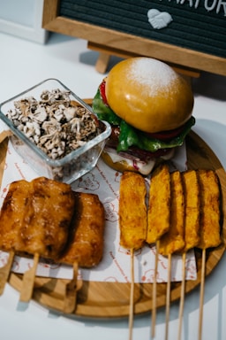A rustic wooden board displaying an assortment of vegan snacks including algae crisps, spicy nuts, garden dip, smoked tofu, miso popcorn, and an edamame bowl.
