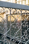 Intricate wrought iron railing with scrolling patterns and heart motifs, set against a blurred background of a glass and metal structure.