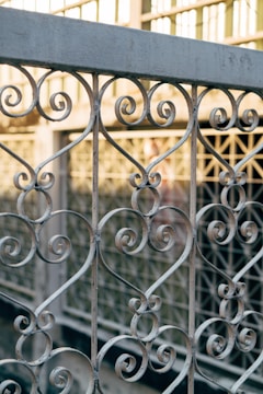 Intricate wrought iron railing with scrolling patterns and heart motifs, set against a blurred background of a glass and metal structure.