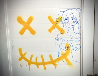 A graffiti drawing with vibrant orange and blue lines on a white panel. The artwork features a blue outline sketch of a cartoonish figure with an expressive face, surrounded by large orange 'X' marks and curved lines resembling a mouth. The bold colors stand out against the plain background.