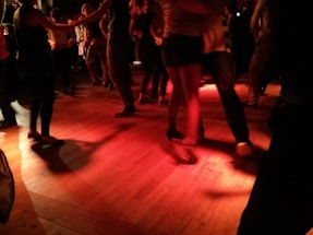 A group of people are dancing in a dimly lit room with red lighting that casts shadows on the wooden floor. The focus is on their legs and feet, with some wearing casual footwear and others in more formal shoes.