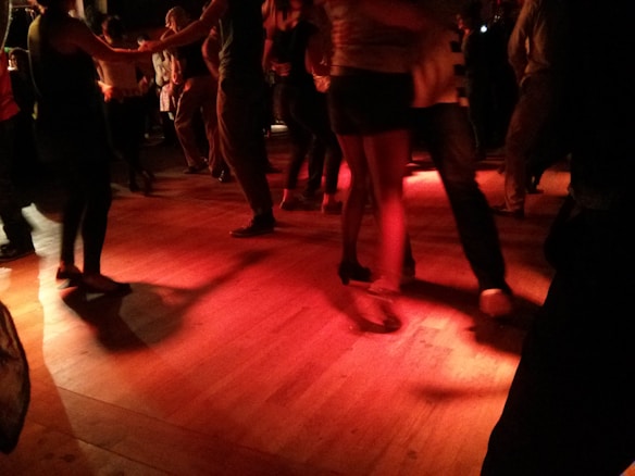 A group of people are dancing in a dimly lit room with red lighting that casts shadows on the wooden floor. The focus is on their legs and feet, with some wearing casual footwear and others in more formal shoes.