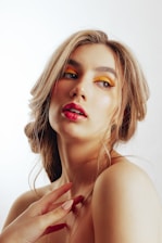 A woman with loose, wavy hair styled in an elegant manner. She has bold, colorful makeup; specifically, yellow eye shadow and bright pink lipstick. Her complexion is fair, with a soft, natural glow. One hand with manicured fingernails is prominently visible near her shoulder, and she gazes slightly to the side with a contemplative expression. The background is plain and light, emphasizing the subject.