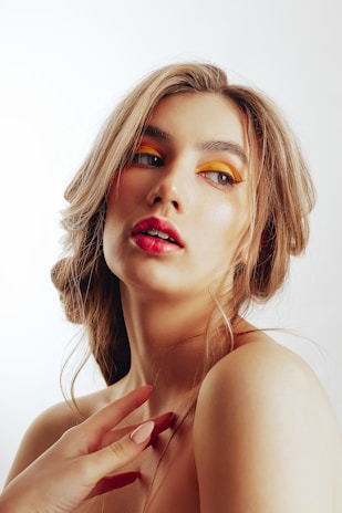 A woman with loose, wavy hair styled in an elegant manner. She has bold, colorful makeup; specifically, yellow eye shadow and bright pink lipstick. Her complexion is fair, with a soft, natural glow. One hand with manicured fingernails is prominently visible near her shoulder, and she gazes slightly to the side with a contemplative expression. The background is plain and light, emphasizing the subject.