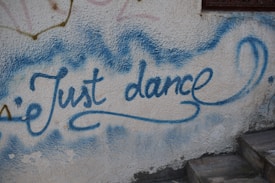 Graffiti art on a textured wall features the words 'Just dance' painted in blue cursive letters. The surrounding area displays additional blue decorative swirls, creating a dynamic and artistic expression.