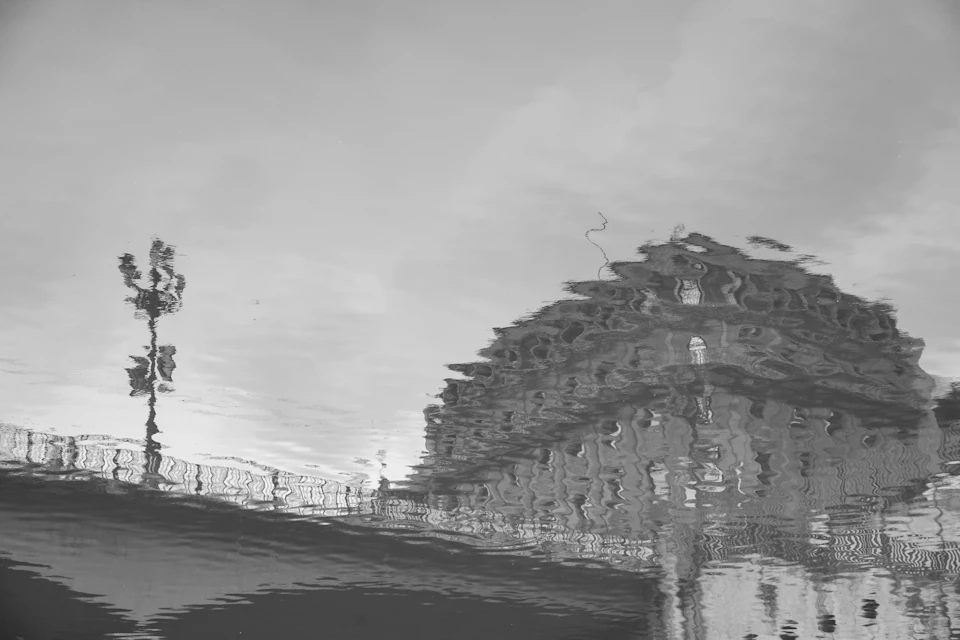 The image depicts a large, ornate building with multiple levels and balconies, situated on a bridge over water. The perspective is from below, creating an interesting reflection effect that adds depth to the scene. A tree can be seen in the foreground, providing a natural element to the urban landscape. The sky above appears hazy, adding a sense of tranquility to the overall composition.