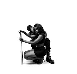 A black and white image featuring a woman crouching while holding a baseball bat, with another person seated slightly behind her. She has curly hair and is dressed in a crop top and skirt, wearing high heels. The person behind is wearing a hat and boots, partially obscured.