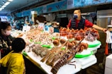 A vibrant seafood market stall displays a variety of fresh crustaceans including lobsters and crabs arranged neatly on ice. Several people, including a vendor and customers wearing masks, engage in transactions at the stall.
