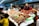A vibrant seafood market stall displays a variety of fresh crustaceans including lobsters and crabs arranged neatly on ice. Several people, including a vendor and customers wearing masks, engage in transactions at the stall.