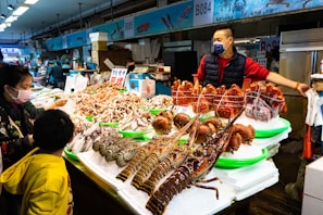 A vibrant seafood market stall displays a variety of fresh crustaceans including lobsters and crabs arranged neatly on ice. Several people, including a vendor and customers wearing masks, engage in transactions at the stall.