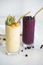 Two tall glasses filled with smoothies are placed side by side on a marble surface. The glass on the left contains a creamy yellow smoothie, garnished with cape gooseberries and green leaves, alongside a gold metal straw. The glass on the right holds a deep purple smoothie, topped with blueberries and also featuring a gold metal straw. Scattered blueberries are visible around the glasses.