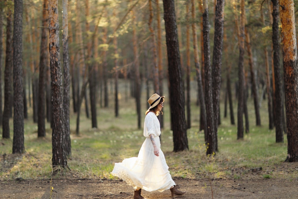 a woman in a white dress and hat walking through a forest