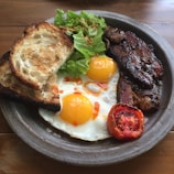 A hearty breakfast plate includes two sunny-side-up eggs with vibrant yellow yolks, crisp bacon strips, a slice of grilled tomato, fresh green lettuce, and two pieces of toasted sourdough bread. The plate is set on a wooden table, creating a rustic and inviting atmosphere.