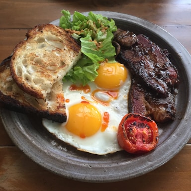 A hearty breakfast plate includes two sunny-side-up eggs with vibrant yellow yolks, crisp bacon strips, a slice of grilled tomato, fresh green lettuce, and two pieces of toasted sourdough bread. The plate is set on a wooden table, creating a rustic and inviting atmosphere.